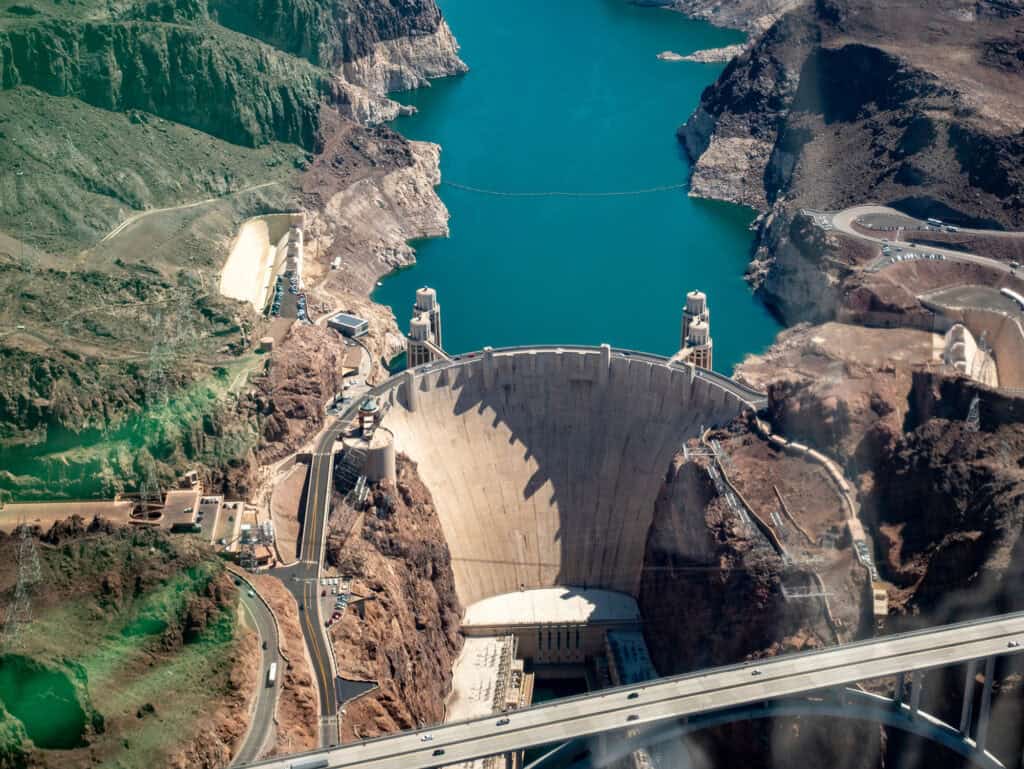 Aerial view of a large concrete dam spanning a canyon with a reservoir behind it. Roads and facilities are visible in the surrounding arid landscape.