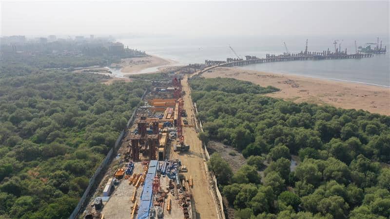 Aerial view of a coastal construction site with heavy machinery surrounded by greenery, a sandy beach, and a pier extending into the sea.