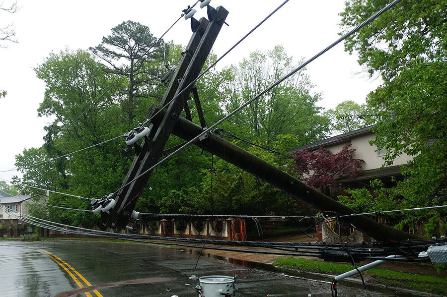 A utility pole is leaning over a wet road with power lines hanging low, possibly due to storm damage; trees and houses are in the background.