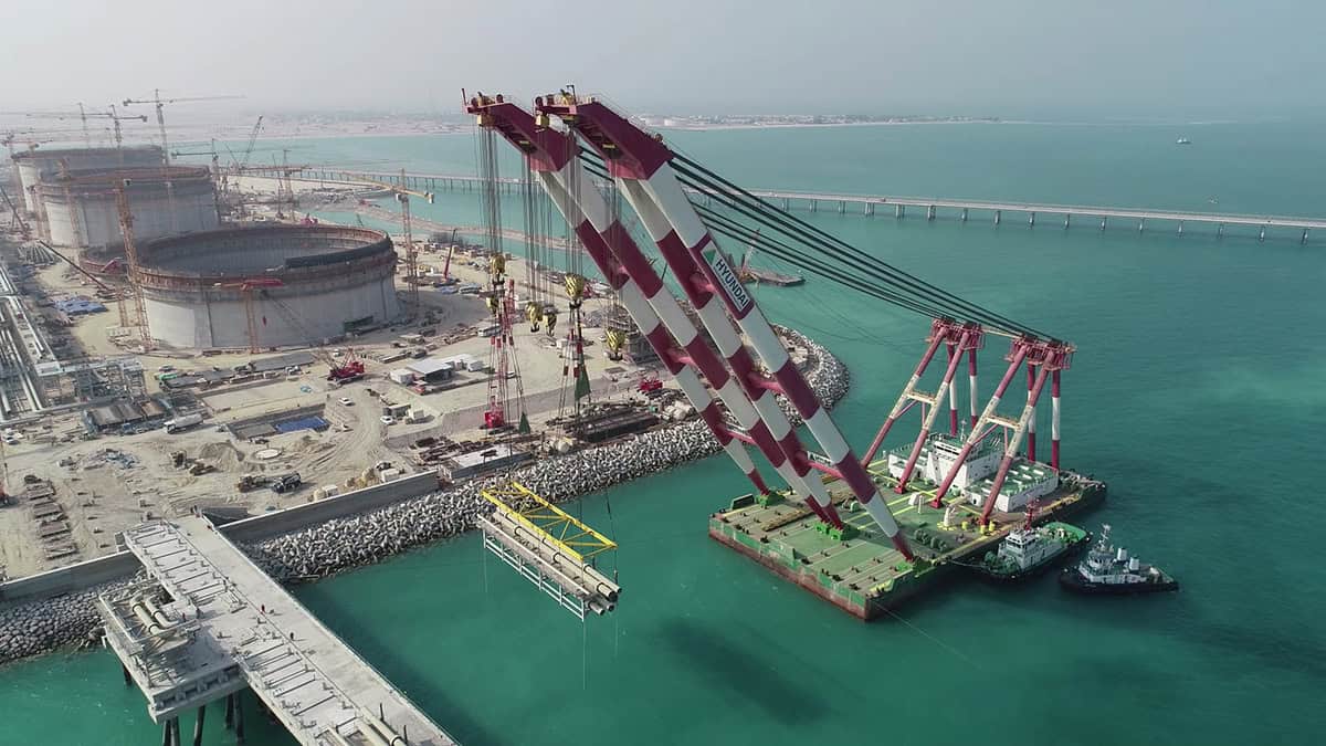 A large crane barge lifts precast concrete structures near storage tanks and a construction site at a waterfront industrial facility. Tugboats assist in the operation.