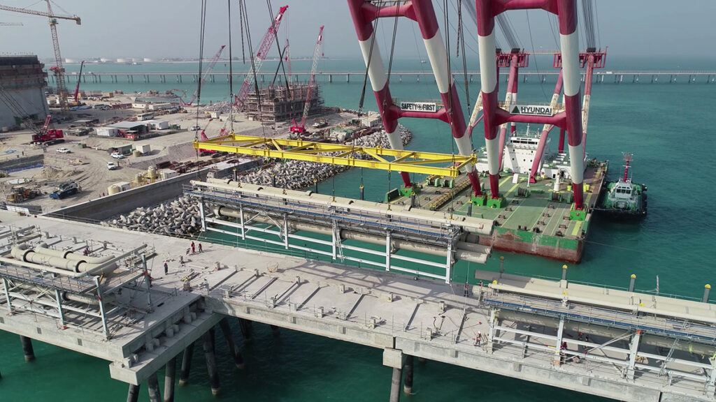 A large crane on a barge lifts a modular concrete structure into place above the water at a construction site, with workers and heavy machinery visible.
