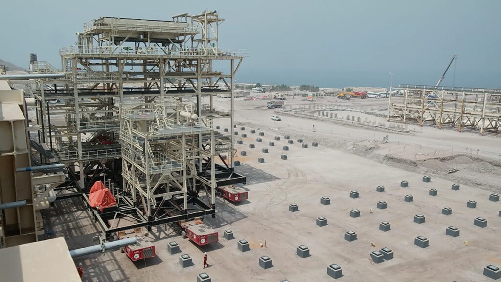 Large industrial construction site with metal framework, scattered precast concrete structures, and a view of the sea in the background under a hazy sky.