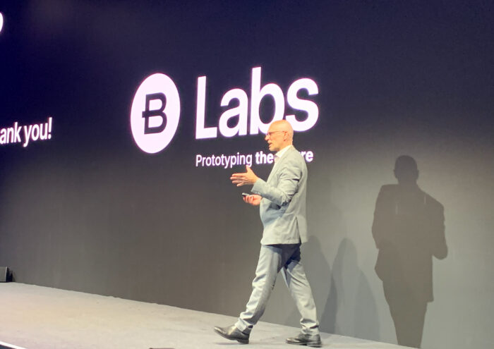 A man in a light gray suit walks on stage in front of a black wall displaying the words “Labs Prototyping the Future” and a logo with a "B" inside a circle.