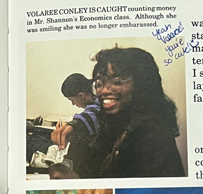 A young woman smiles at the camera while counting money in a classroom. A handwritten note says, "Yeah Volare! so cute!" near the photo. Someone is seated behind her.