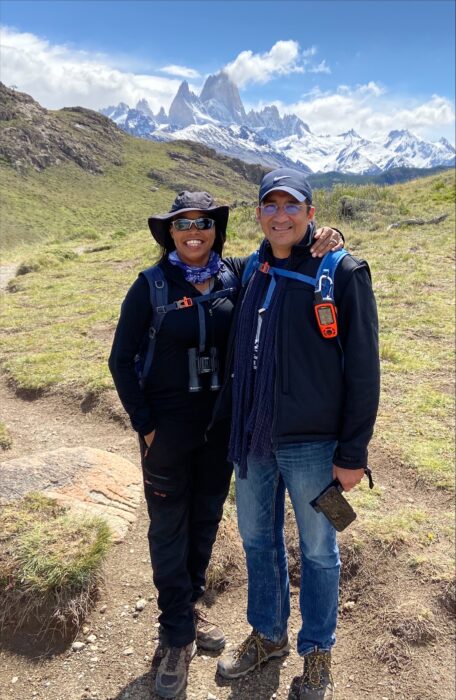 Two people dressed in hiking gear stand smiling on a grassy trail with snow-capped mountains in the background under a partly cloudy sky.