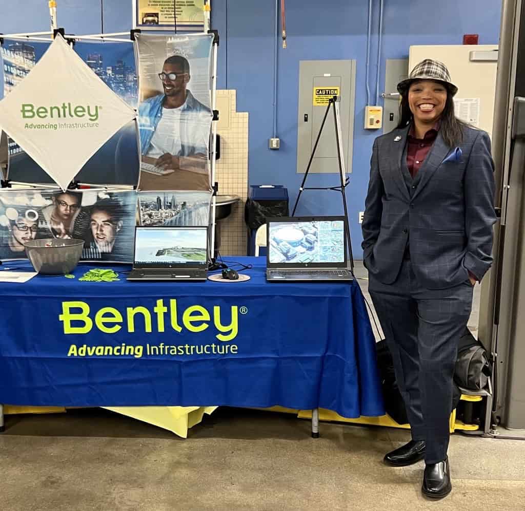A person in a suit stands beside a Bentley Advancing Infrastructure booth with two laptops and promotional materials on the table.
