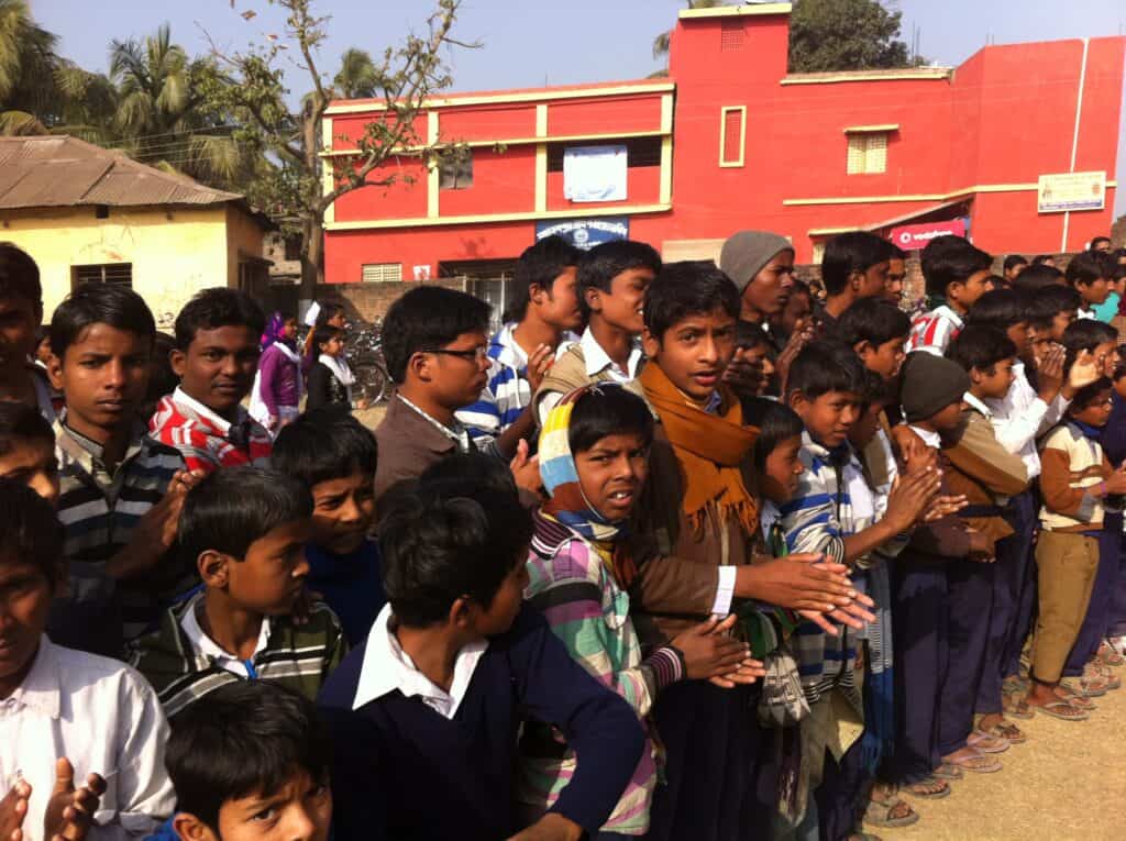 A group of school-age children stand outdoors in rows, some clapping, with buildings and trees in the background under sunlight—echoing Apurba Tribedi’s vision of engineering for impact in communities.