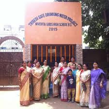 A group of women and a boy stand in front of a building labeled "Trivedi Safe Drinking Water Project Nivedita Girls High School (HS) 2015," highlighting an engineering effort led by Apurba Tribedi.
