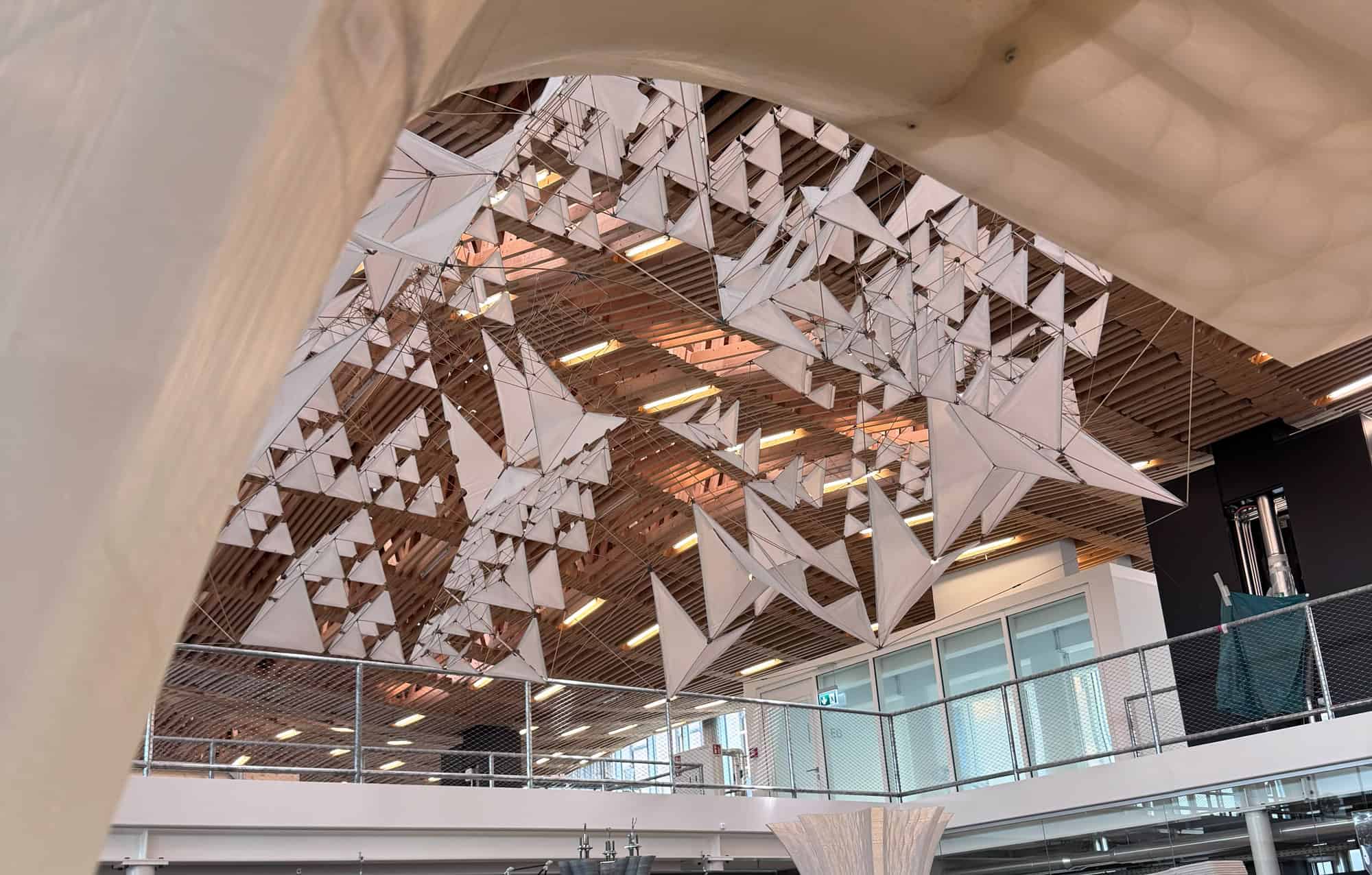 Geometric white origami-like sculptures hang from the ceiling in a modern building with wooden slats and glass-panel walls.