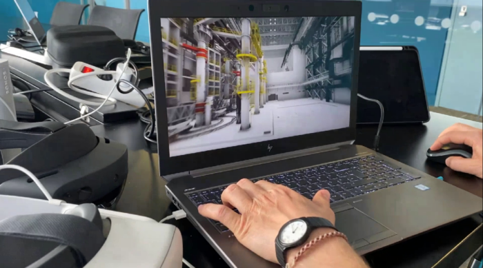 A person operates a laptop showing a 3D rendering of industrial equipment, surrounded by various electronic devices on a desk.