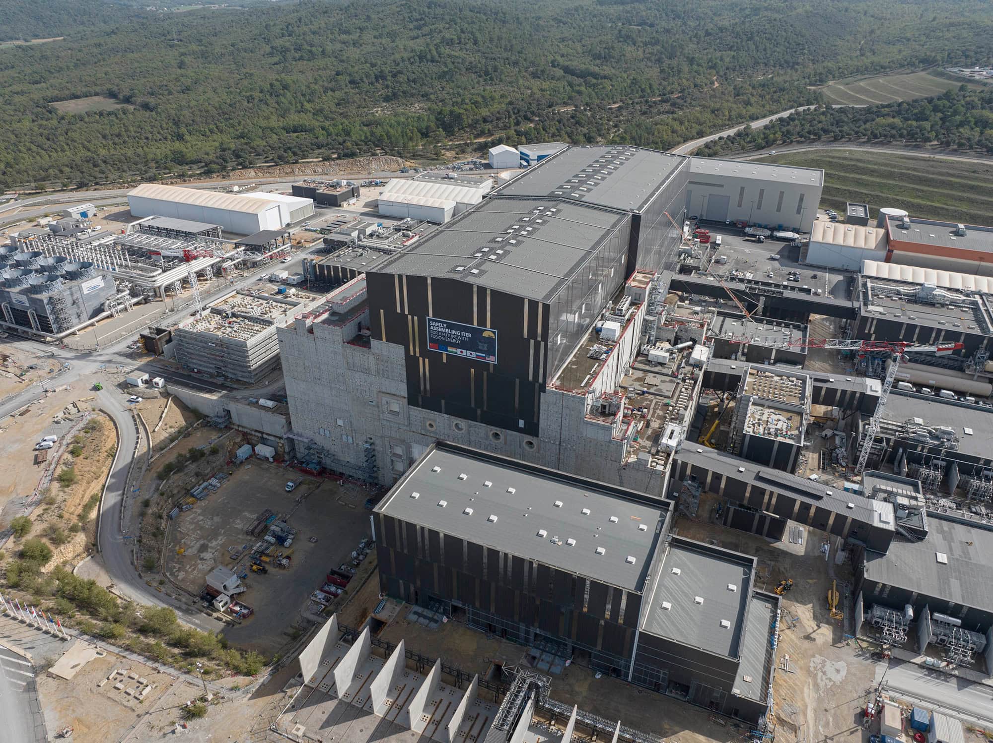 Aerial view of a large industrial facility under construction, surrounded by forested areas and roads. Several buildings and cranes are visible, indicating ongoing development work.