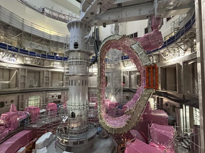 Industrial interior with scaffolding and machinery, featuring a large, circular metallic structure in the center surrounded by pink protective coverings.