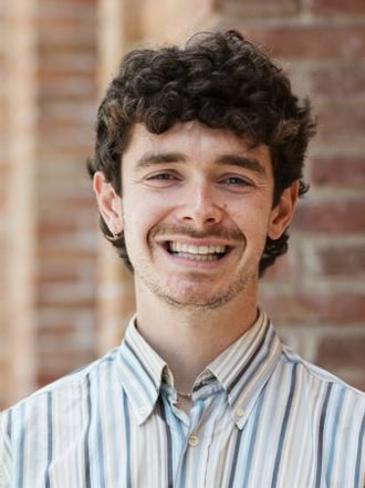 A young man with curly brown hair and a striped shirt smiles while standing in front of a brick wall.