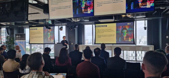 A speaker presents a slide titled "What is Open Data?" to an audience in a modern conference room with large windows and city views.