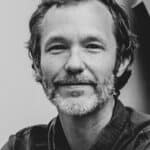 Black and white portrait of a middle-aged man with a beard and slightly tousled hair, wearing a button-up shirt and looking directly at the camera.