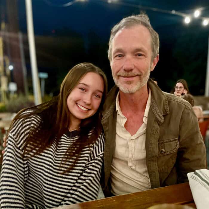 A smiling young woman in a striped sweater sits beside a smiling man in a jacket at an outdoor table, with people and lights in the background.