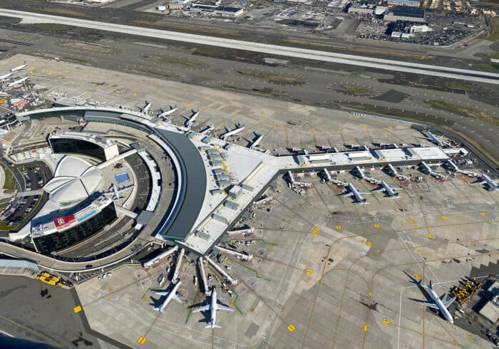 Aerial view of a large airport terminal with multiple airplanes parked at gates and several runways and taxiways visible.