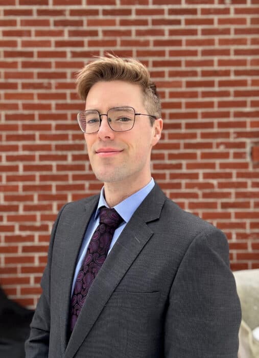 Person in a suit with glasses standing in front of a brick wall.