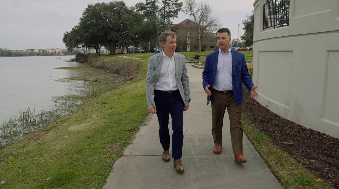 Two men in jackets walk and talk on a lakeside path with trees and buildings in the background.