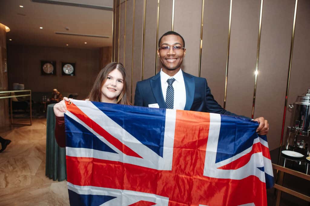 Two people standing indoors, dressed formally, holding a large Union Jack flag between them.