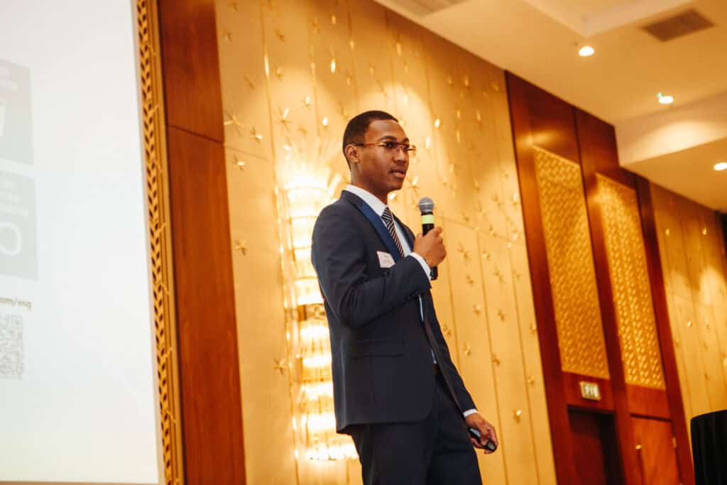 A man in a suit speaks into a microphone while standing on stage in a conference room with wood-paneled walls and a projection screen.
