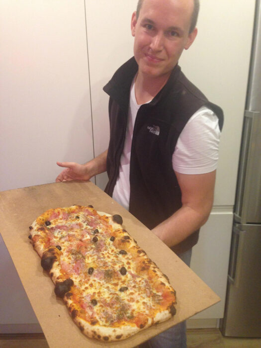 Bentley CTO Julien Moutte holding a rectangular pizza on a wooden board in a kitchen.