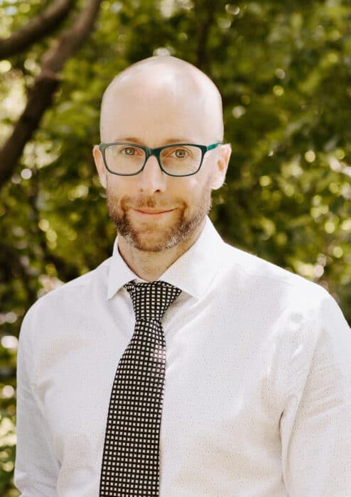 A man with a bald head, glasses, and a beard wears a white shirt and patterned tie, standing outdoors with green foliage in the background.