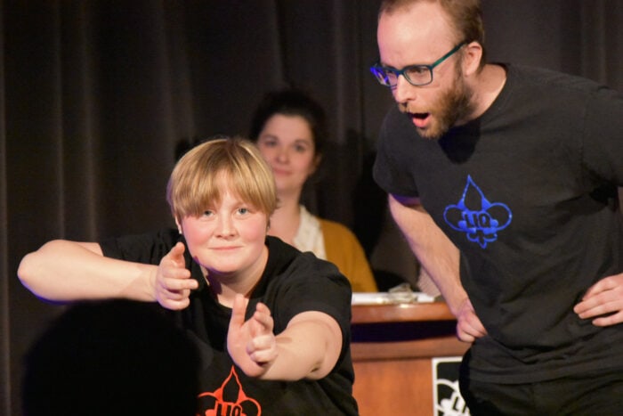 Two people wearing black shirts perform on stage; one points finger guns forward while the other reacts animatedly. A seated person watches from behind.