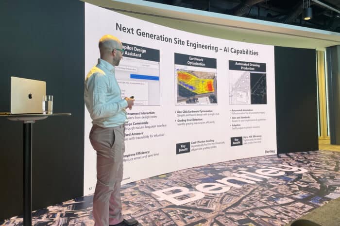 A man stands next to a presentation screen displaying slides on AI capabilities in next generation site engineering, including design assistance, earthwork optimization, and drawing production.