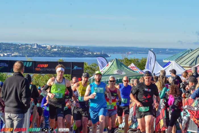 A group of runners participate in an outdoor race, surrounded by spectators and tents, with a scenic city and water view in the background.