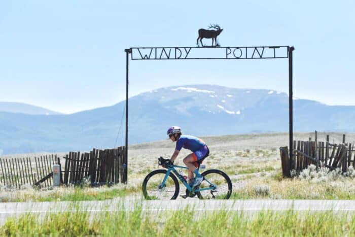 A cyclist rides past a sign reading "Windy Point" with a deer silhouette on top. Snow-capped mountains and a wooden fence are visible in the background.