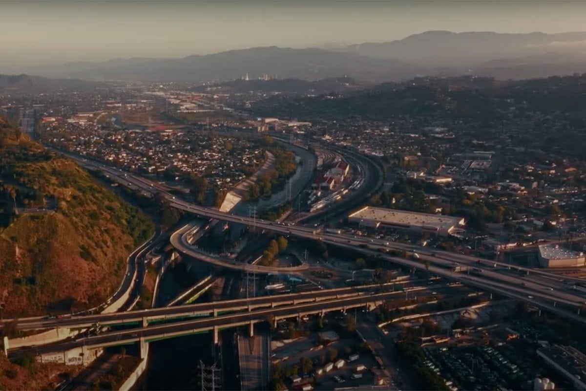 Aerial view of a sprawling freeway interchange, a testament to exceptional infrastructure design, surrounded by urban buildings, neighborhoods, and hills, with mountains visible in the background under a hazy sky.