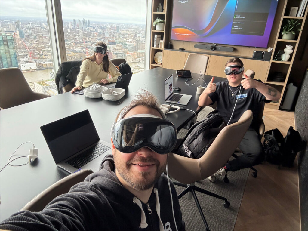 Three people sit around a conference table with laptops, wearing large black goggles, in a high-rise office with a city view.