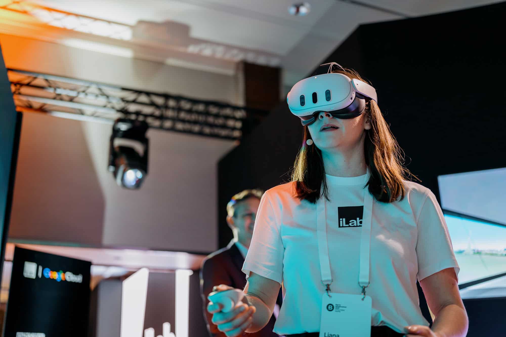 Liana O'Cleirigh wearing a white VR headset and holding controllers stands indoors, participating in a virtual reality experience at a tech event.