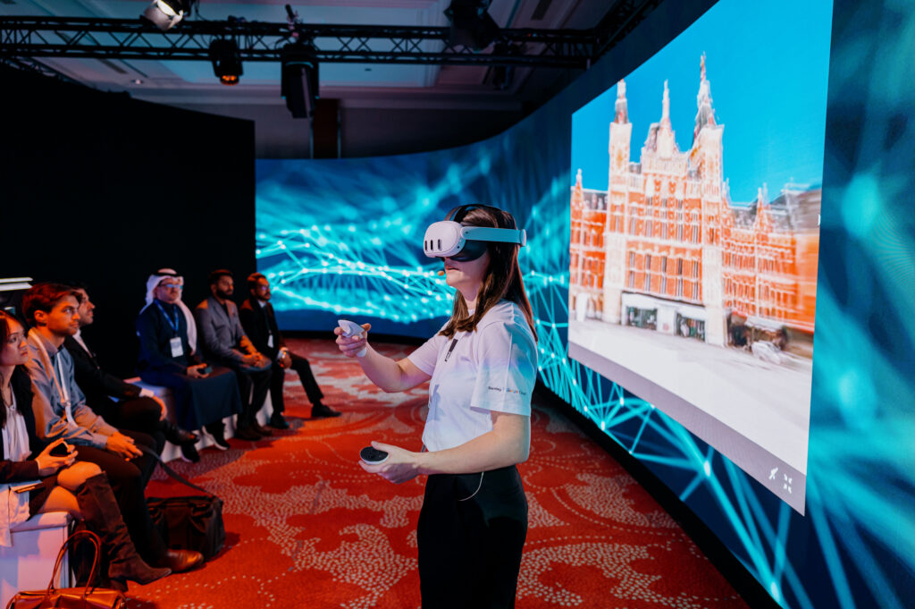 Liana O'Cleirigh wearing a VR headset stands and interacts with a controller in front of an audience, with a large screen displaying a digital image of a building in the background.