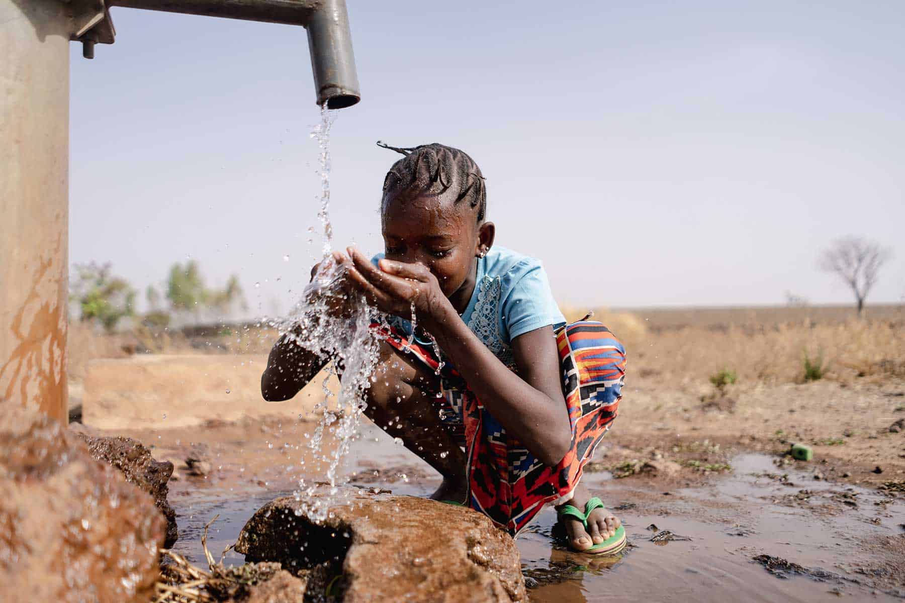 A child crouches by a pipe, drinking water with hands cupped—an image of hope and resilience in an outdoor, rural setting with dry grass and scattered trees, showing the power of survival on our changing planet.