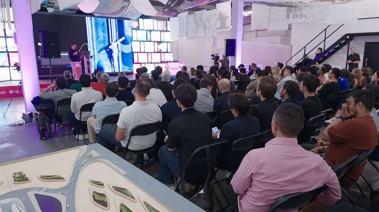 A group of people sit facing a speaker presenting on stage in a modern, open room with screens and architectural models.