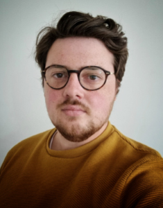 A person with short brown hair, a beard, and glasses is wearing a mustard yellow sweater and standing in front of a plain light-colored background.