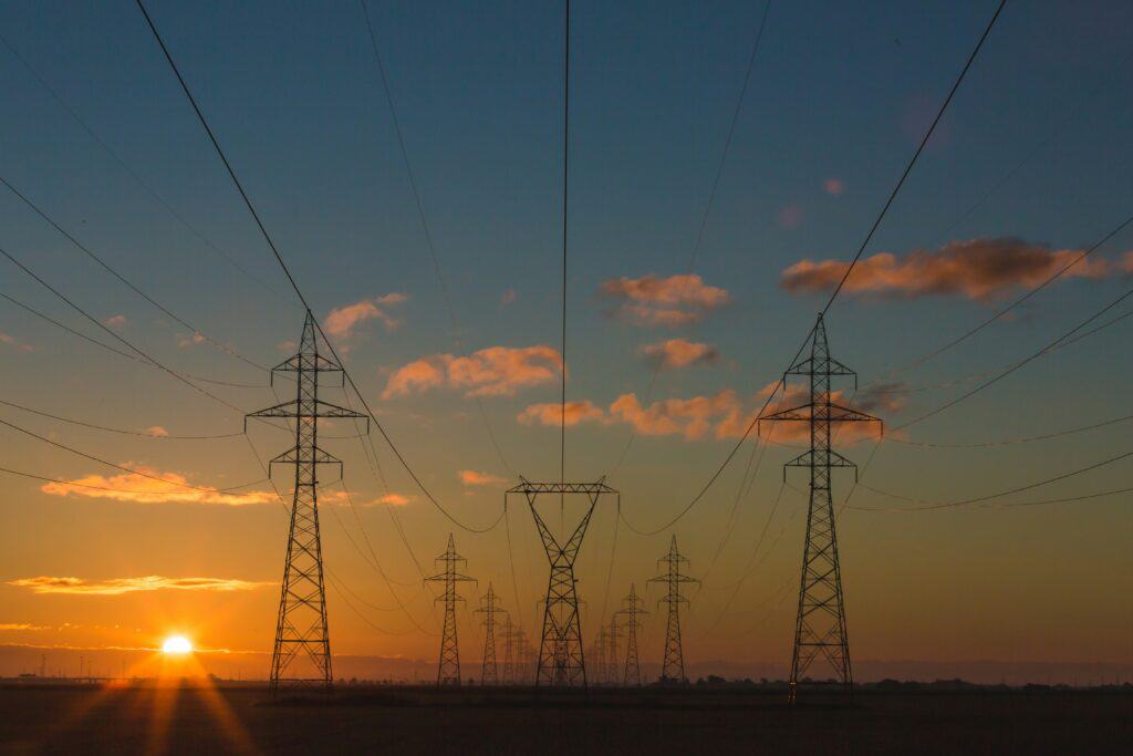 Power lines and transmission towers stretch across a field at sunset, with the sun low on the horizon and clouds scattered in the sky.