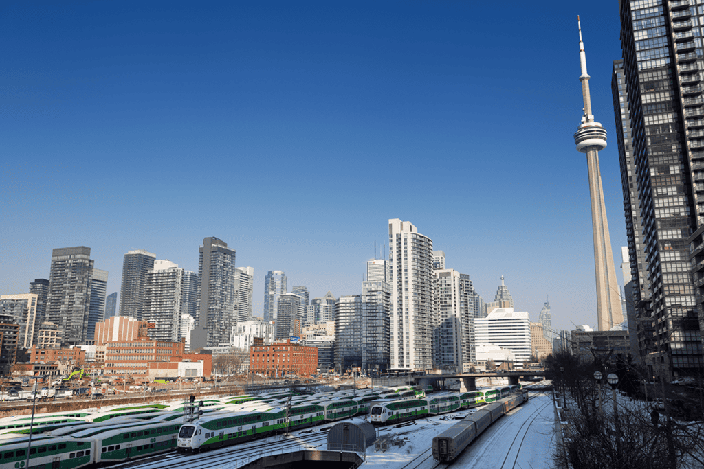 Toronto city skyline with modern high-rise buildings, the CN Tower, and trains on Toronto rail tracks in the foreground on a clear day.