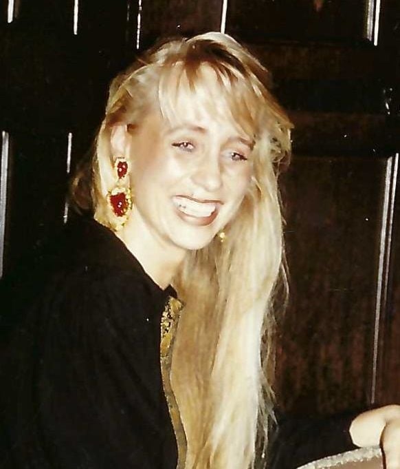 A woman with long blonde hair smiles, wearing large red earrings and a black top, sitting in front of a dark wood background. Her style hasn’t changed much over the years, remaining timeless and elegant.