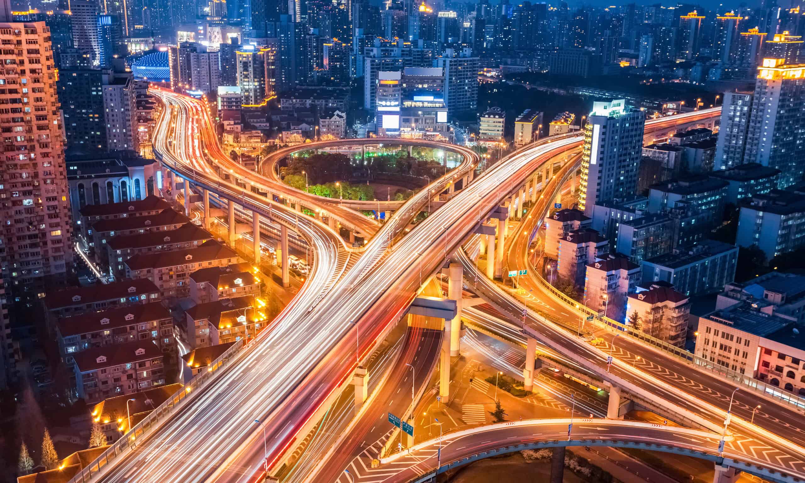 Aerial view of a busy urban highway interchange at night, with light trails from vehicles and tall buildings surrounding the roads.