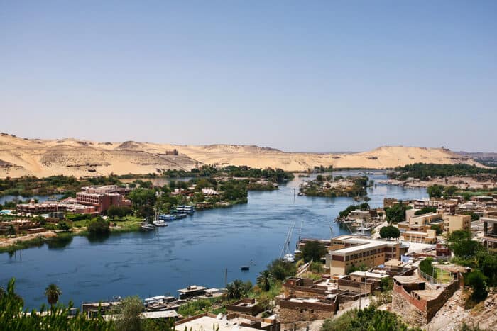 A scenic view of a river winding through a desert landscape with buildings and boats on its banks, surrounded by lush vegetation and sand dunes under a clear blue sky.