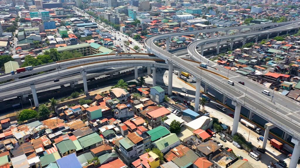 Aerial view of a multi-level highway interchange, part of tomorrow’s roads, above a densely populated urban area with colorful rooftops and various vehicles navigating the digital infrastructure below.