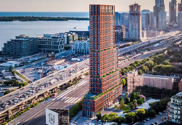 A tall, uniquely tiered red-brick high-rise building stands near a highway and the busiest rail corridor, with cityscape and waterfront in the background.