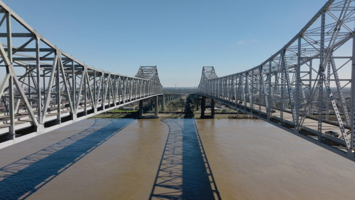 Twin steel bridges span across a wide, brown river under a clear blue sky, with their reflections visible in the water below.