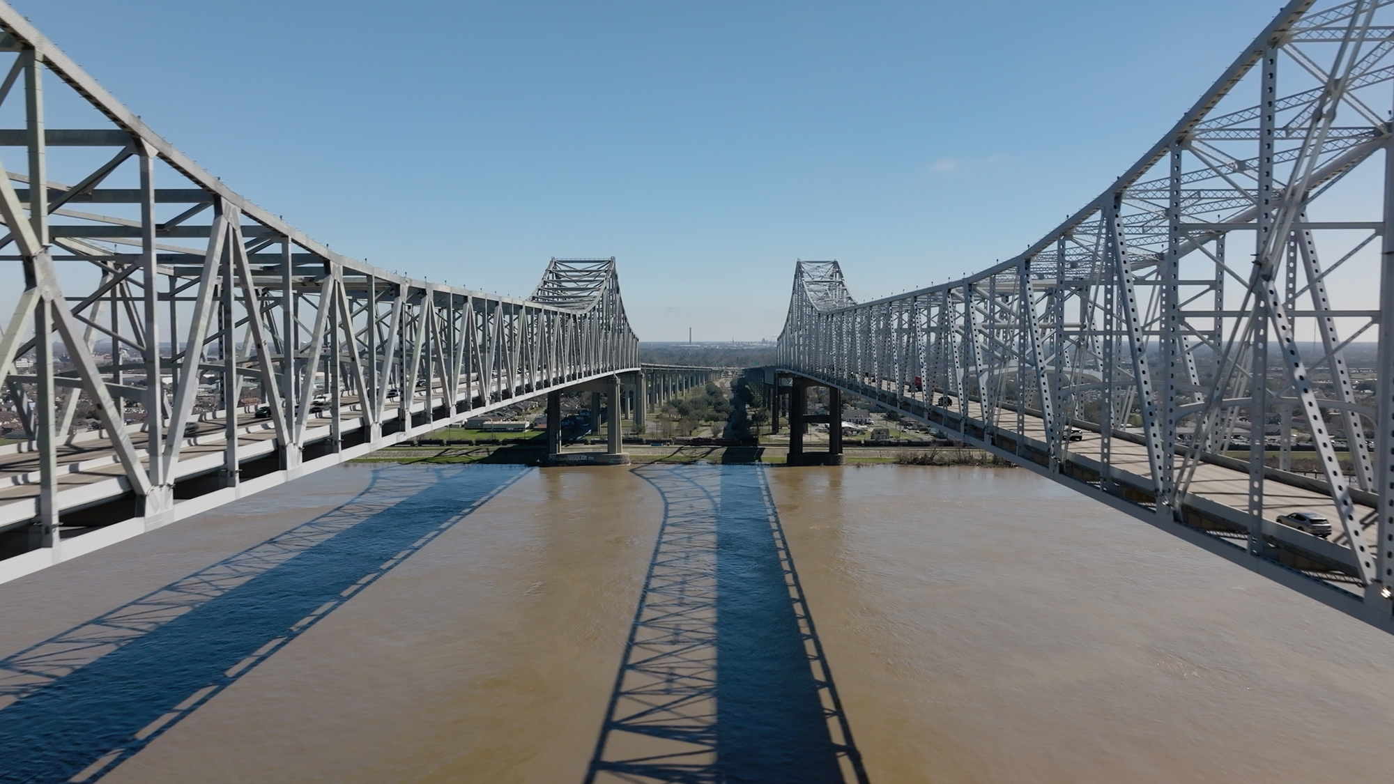 Twin steel bridges span across a wide, brown river under a clear blue sky, with their reflections visible in the water below.