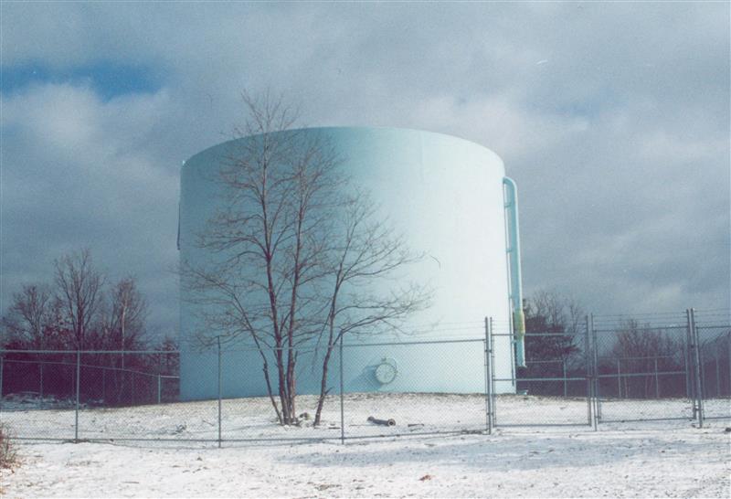 A large cylindrical water tank, serving as a foundation for success in resource management, stands encircled by a chain-link fence. This scene unfolds in a snowy landscape dotted with bare trees, embodying the careful planning necessary for thriving in challenging environments.