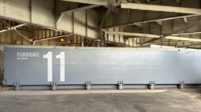 A large, gray floodgate labeled "FLOODGATE MF01-011" and the number "11" stands beneath a metal bridge structure.