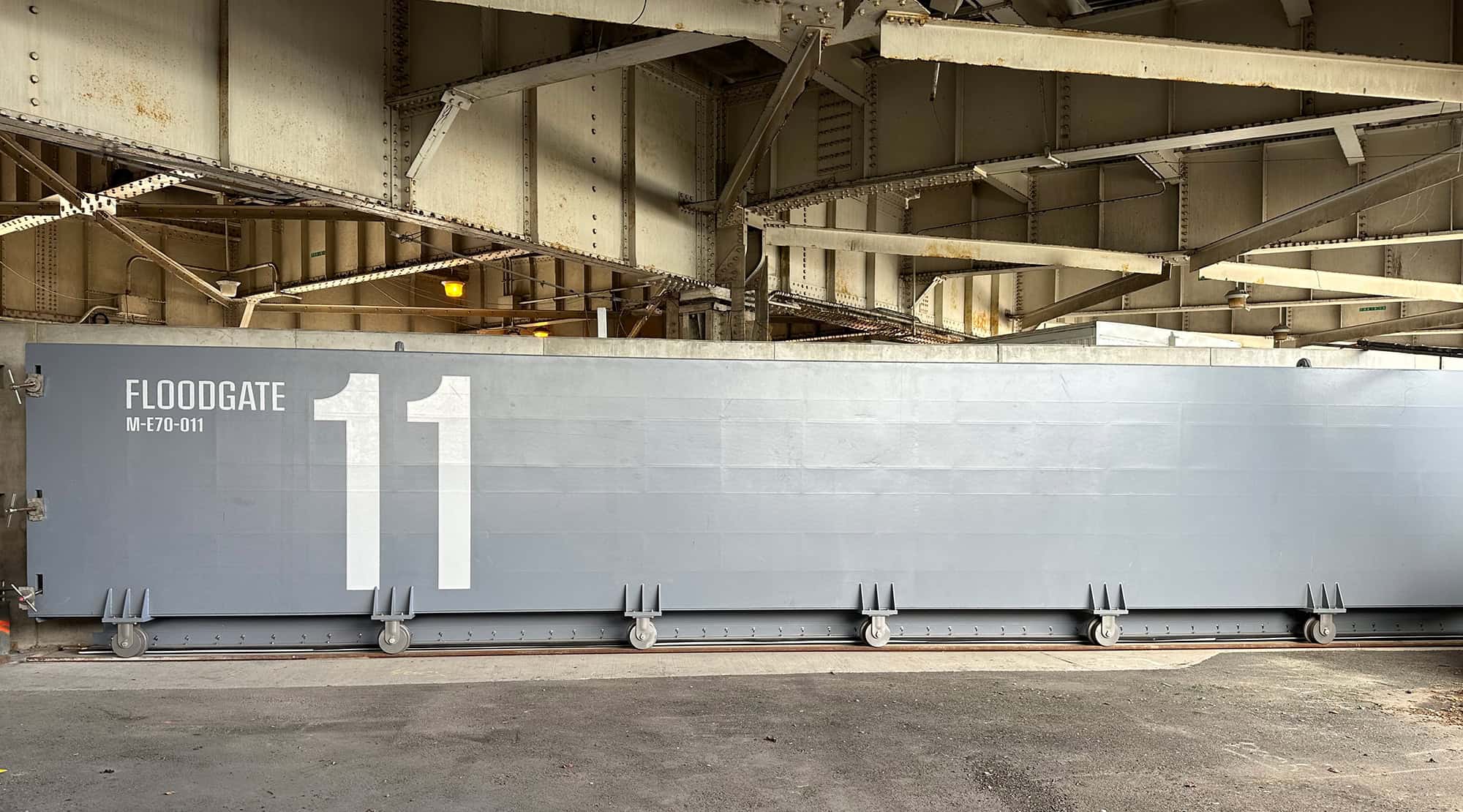 A large, gray floodgate labeled "FLOODGATE MF01-011" and the number "11" stands beneath a metal bridge structure.